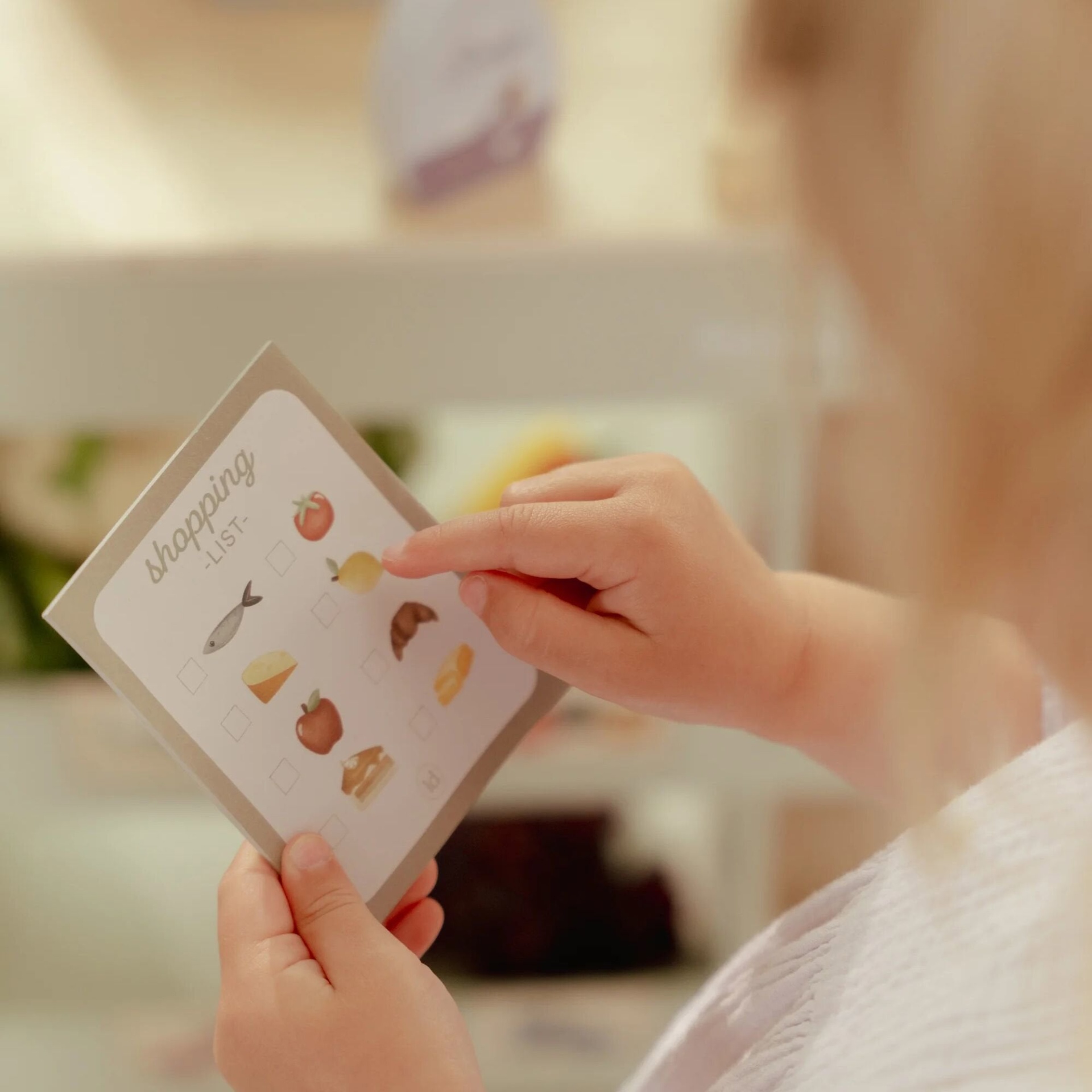 Lista de compras em papel com ícones de alimentos e mãos infantis segurando-a