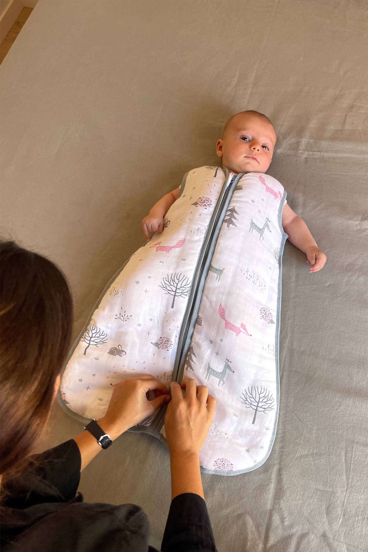 Bebé deitado a usar saco de dormir branco com padrão em cinza e rosa, com fecho de correr cinzento sendo fechado por pessoa.
