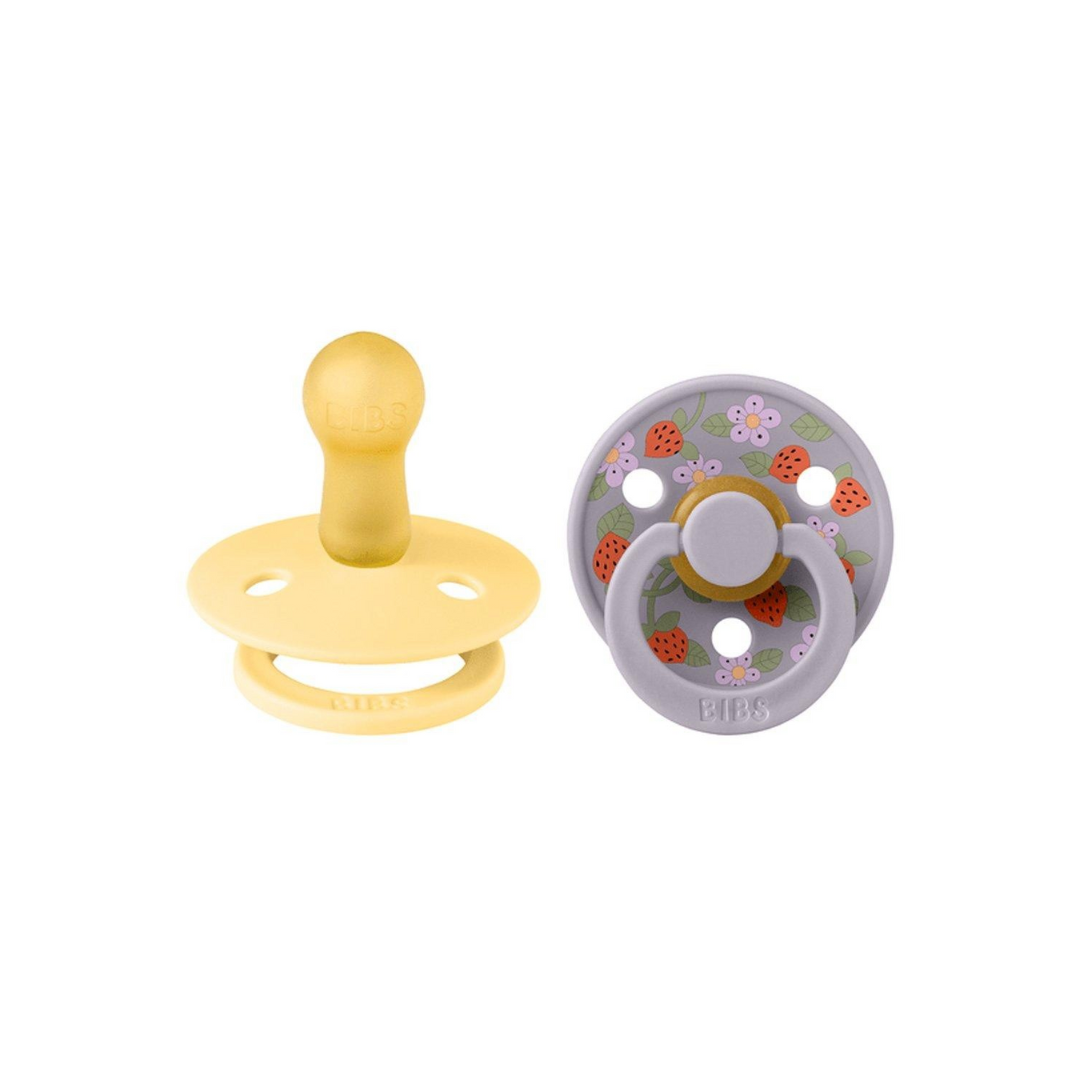 Dois chupetas de bebé, uma amarela lisa e outra cinzenta com padrão de morangos e flores.