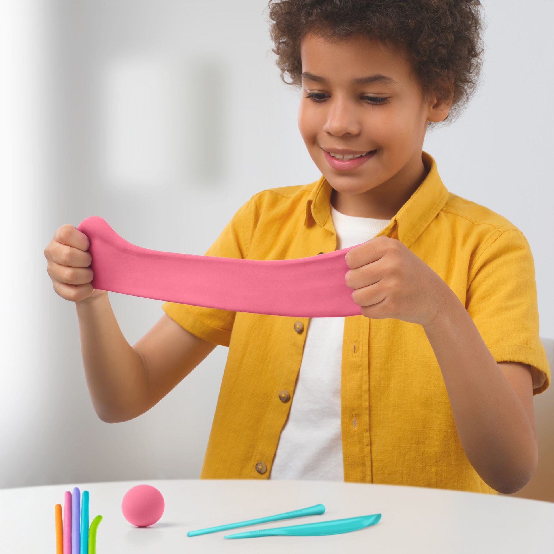 Criança a esticar massa de modelar cor-de-rosa com ferramentas de modelagem numa mesa branca