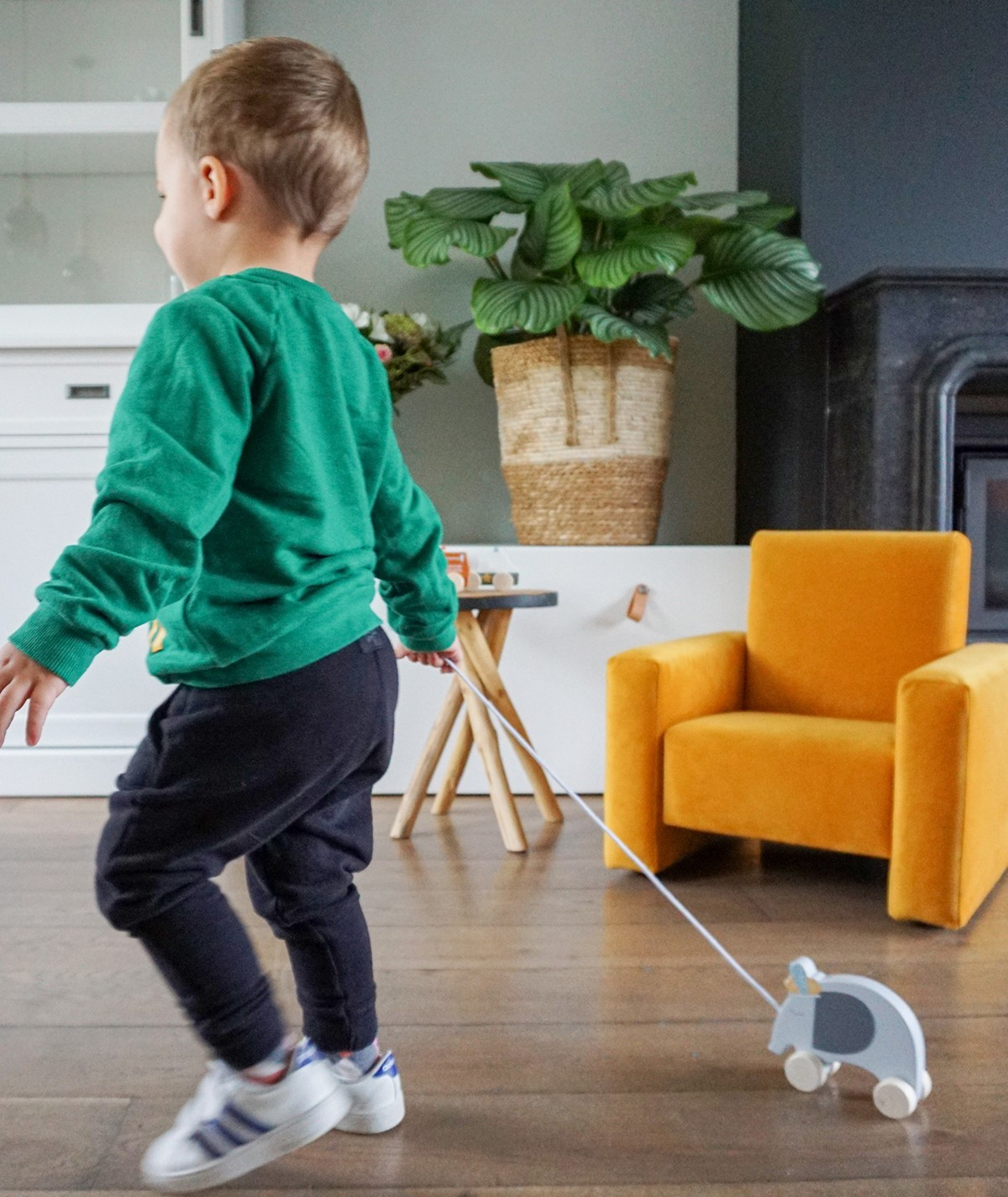 Criança a brincar com brinquedo de madeira puxado por corda em sala com cadeira amarela.