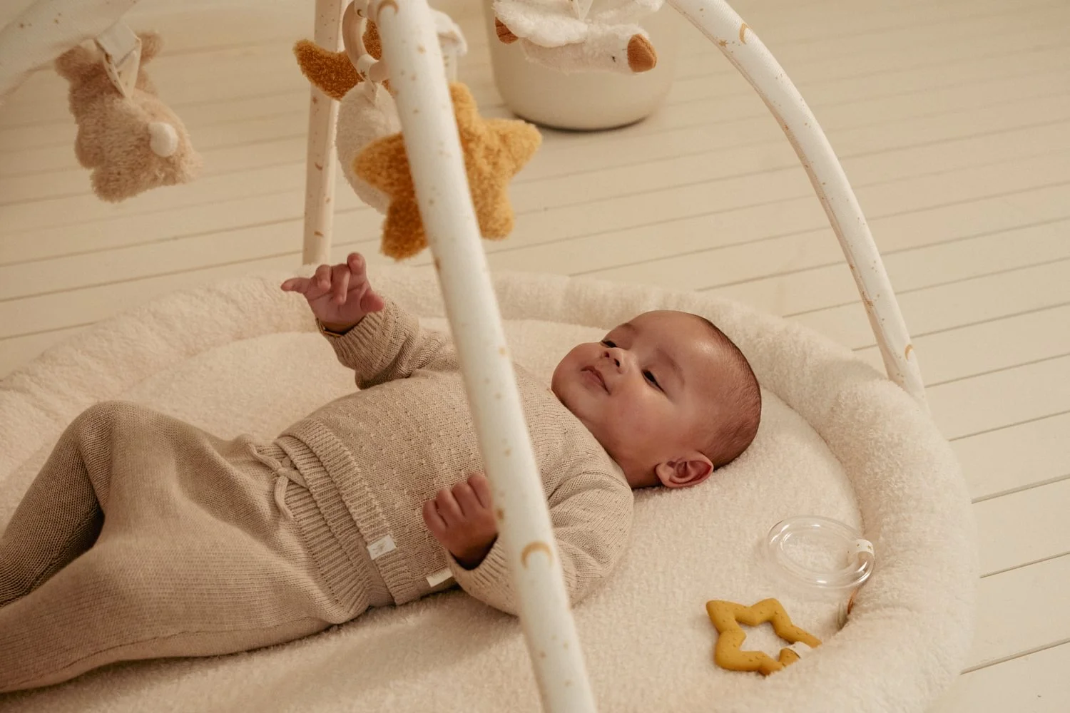 Tapete de atividades para bebé creme com brinquedos pendurados e bebé vestido de malha bege.