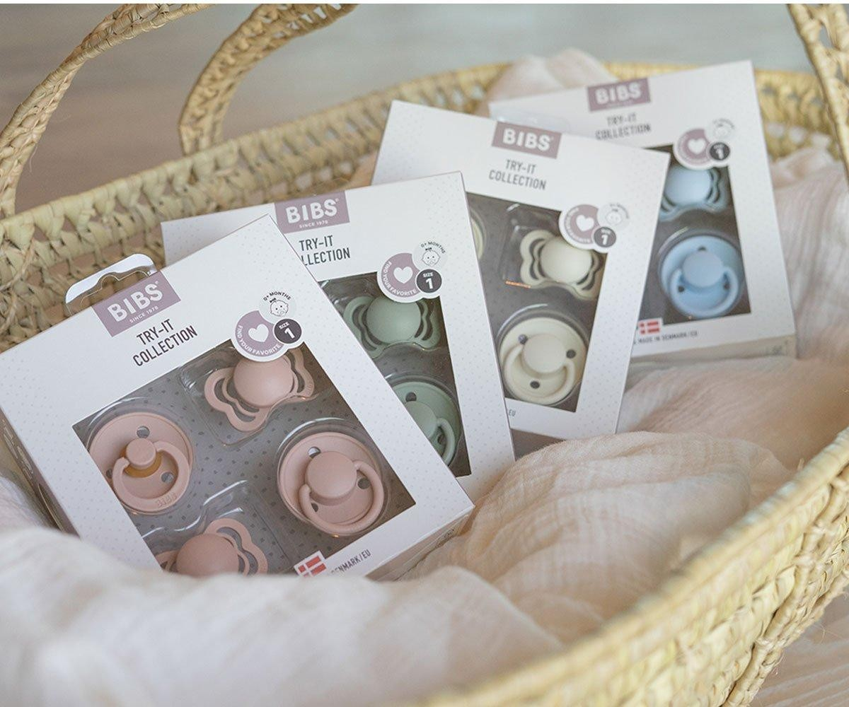 Embalagens de chupetas BIBS TRY-IT COLLECTION de cores rosa, verde, bege e azul numa cesta de palha.