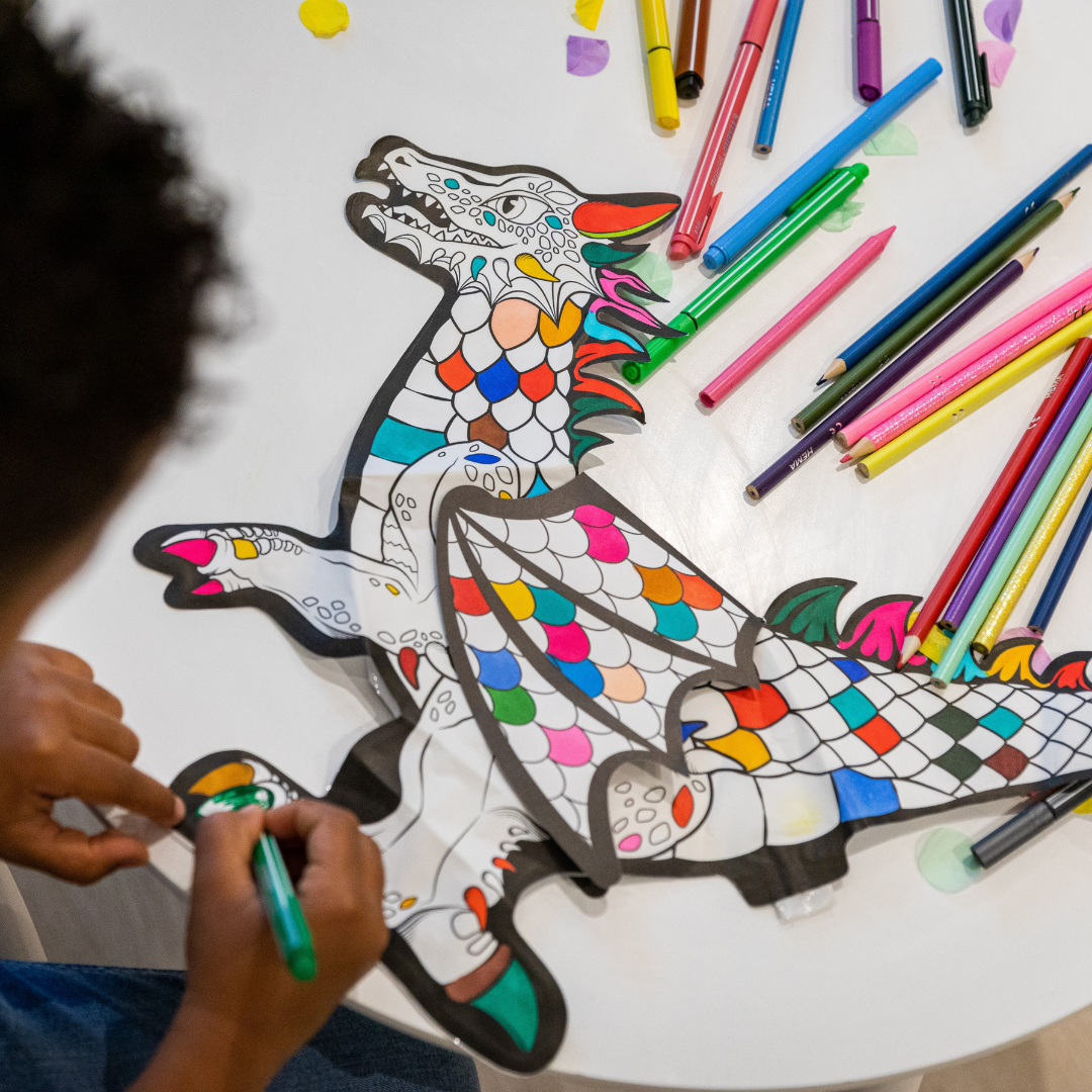 Dragão de papel para colorir com canetas coloridas numa mesa branca, criança a pintar.