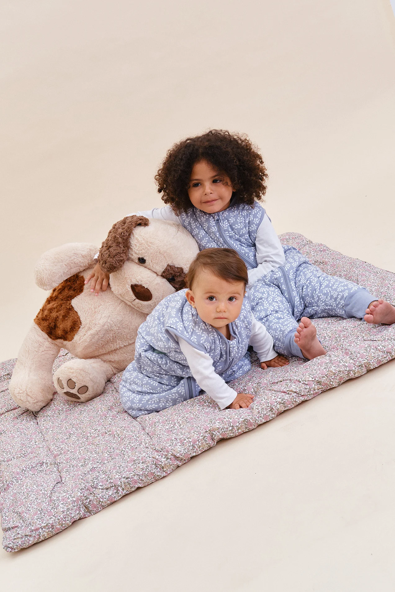 Duas crianças com roupa de dormir azul clara estampada, sentadas num edredão floral rosa com urso de peluche castanho e branco.