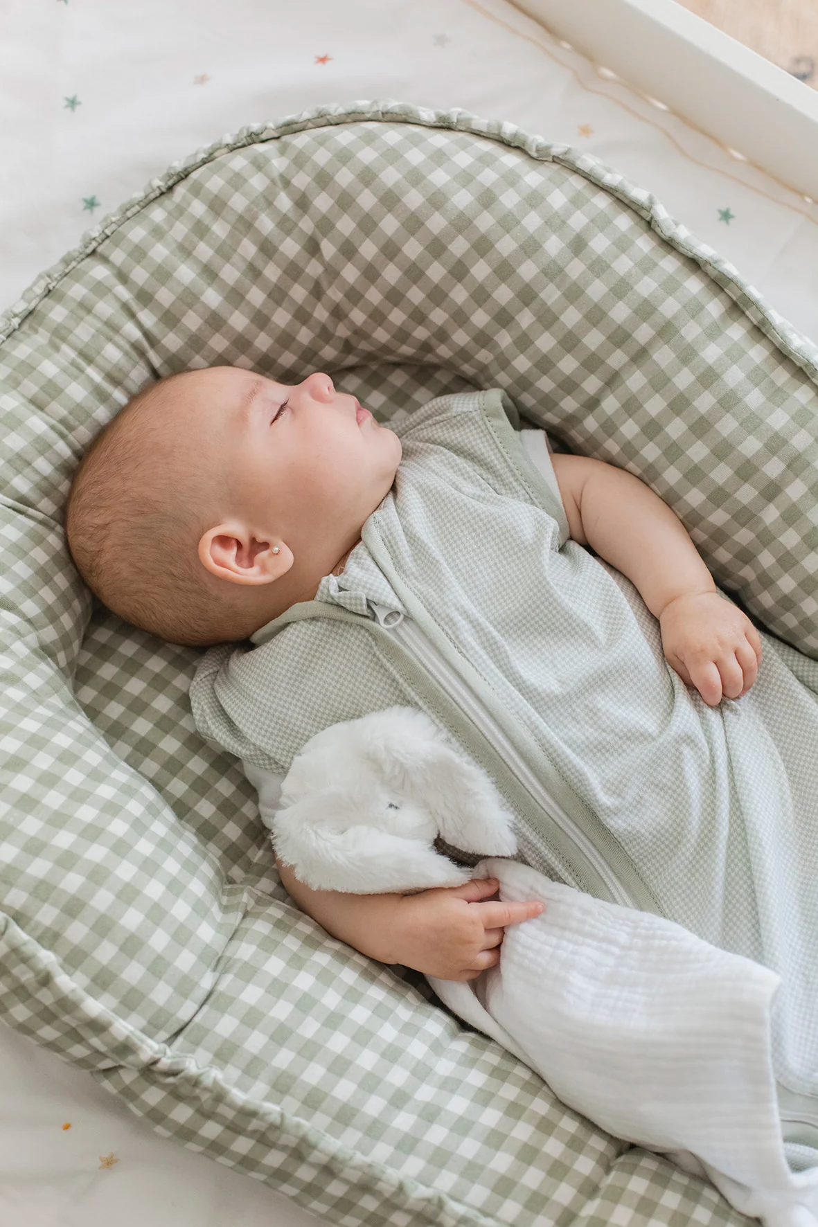 Bebé adormecido numa cama de tecido xadrez verde e branco com saco de dormir cinzento claro e brinquedo de peluche branco