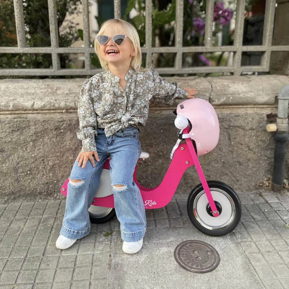 Menina loira sentada numa mini moto rosa de criança com capacete cor de rosa, vestindo camisa floral e jeans rasgados