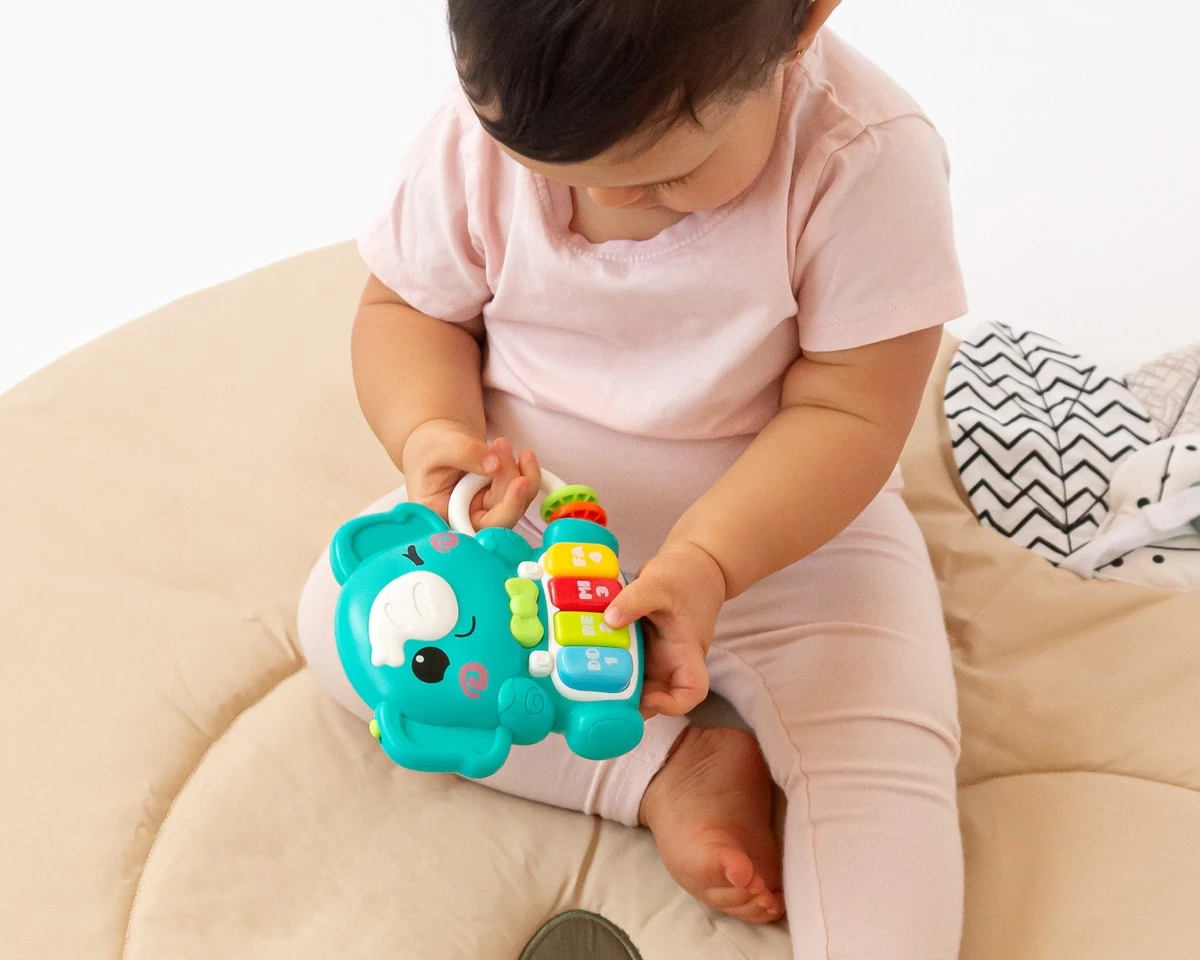 Bebé com roupa rosa a segurar brinquedo musical azul em forma de elefante com teclas coloridas.