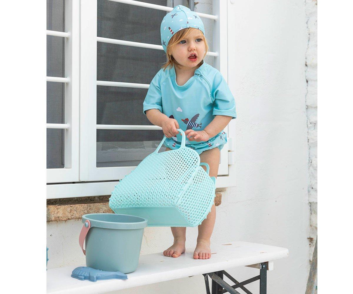 Criança vestindo roupas azuis segurando bolsa azul em bancada branca perto de porta com vidros.