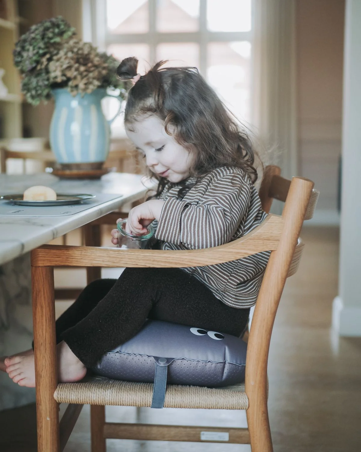 Criança sentada em cadeira de madeira clara com almofada cinza com olhos desenhados