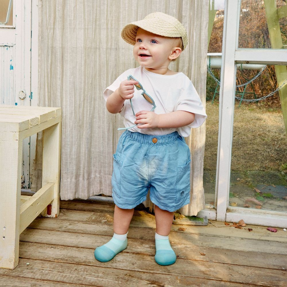 Criança com roupa casual clara e boné bege em chão de madeira junto a cortina e porta de vidro