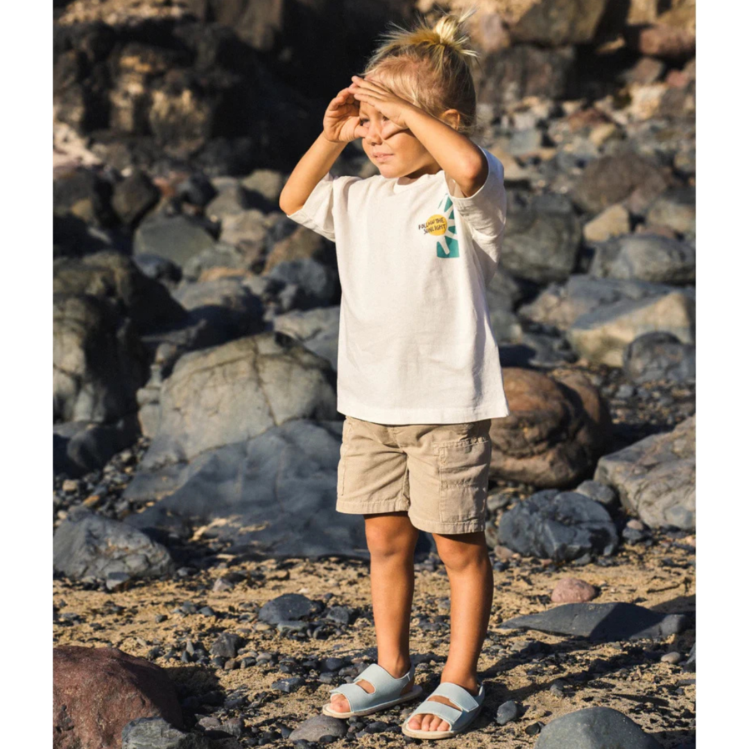 Criança de pé em terreno rochoso com roupa clara e sandálias.