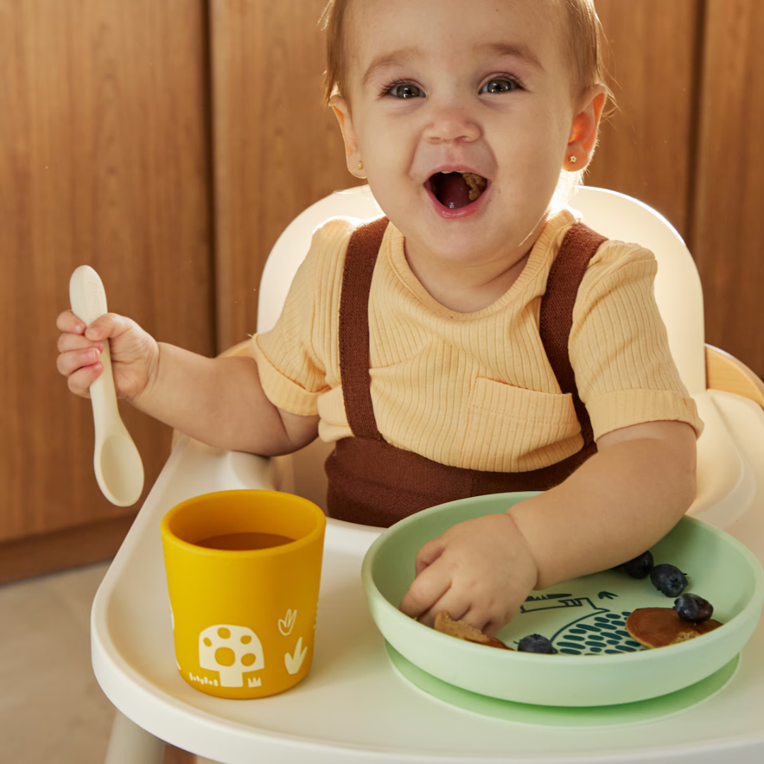 Criança sorridente com colher branca, copo amarelo e prato verde com comida sentada em cadeirão branco
