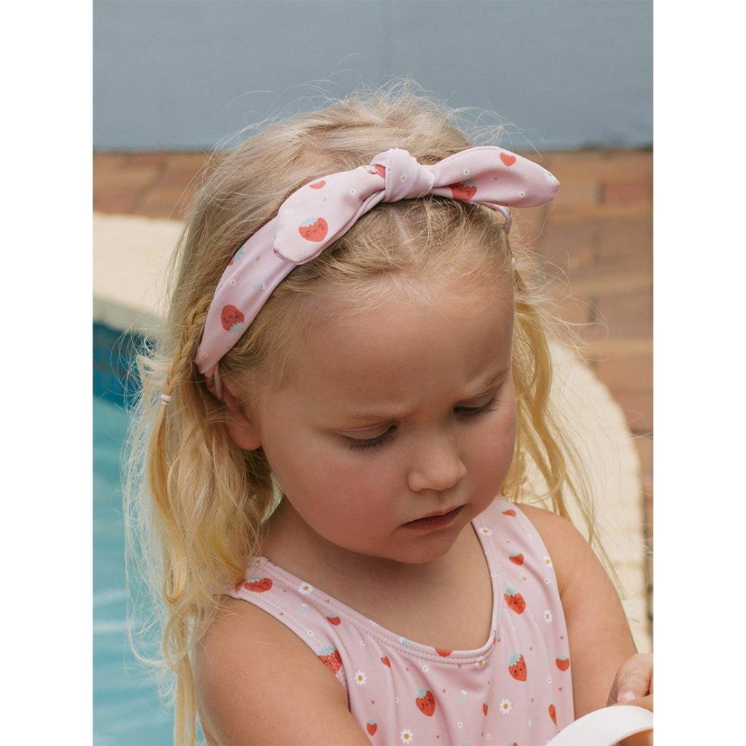 Menina loira com fato de banho e faixa de cabelo cor-de-rosa com morangos perto de piscina