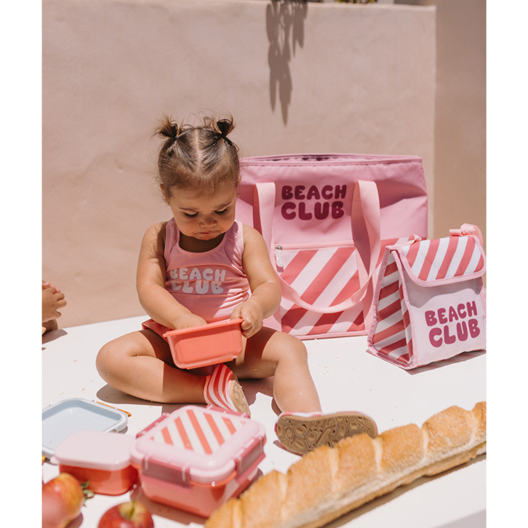 Criança com fato de banho rosa BEACH CLUB e embalagens de almoço cor-de-rosa com padrão às riscas BEACH CLUB numa mesa com pão e maçãs.