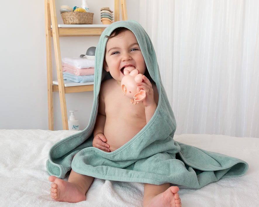 Bebé sentado com toalha verde água com capuz e biberão cor de rosa numa cama branca