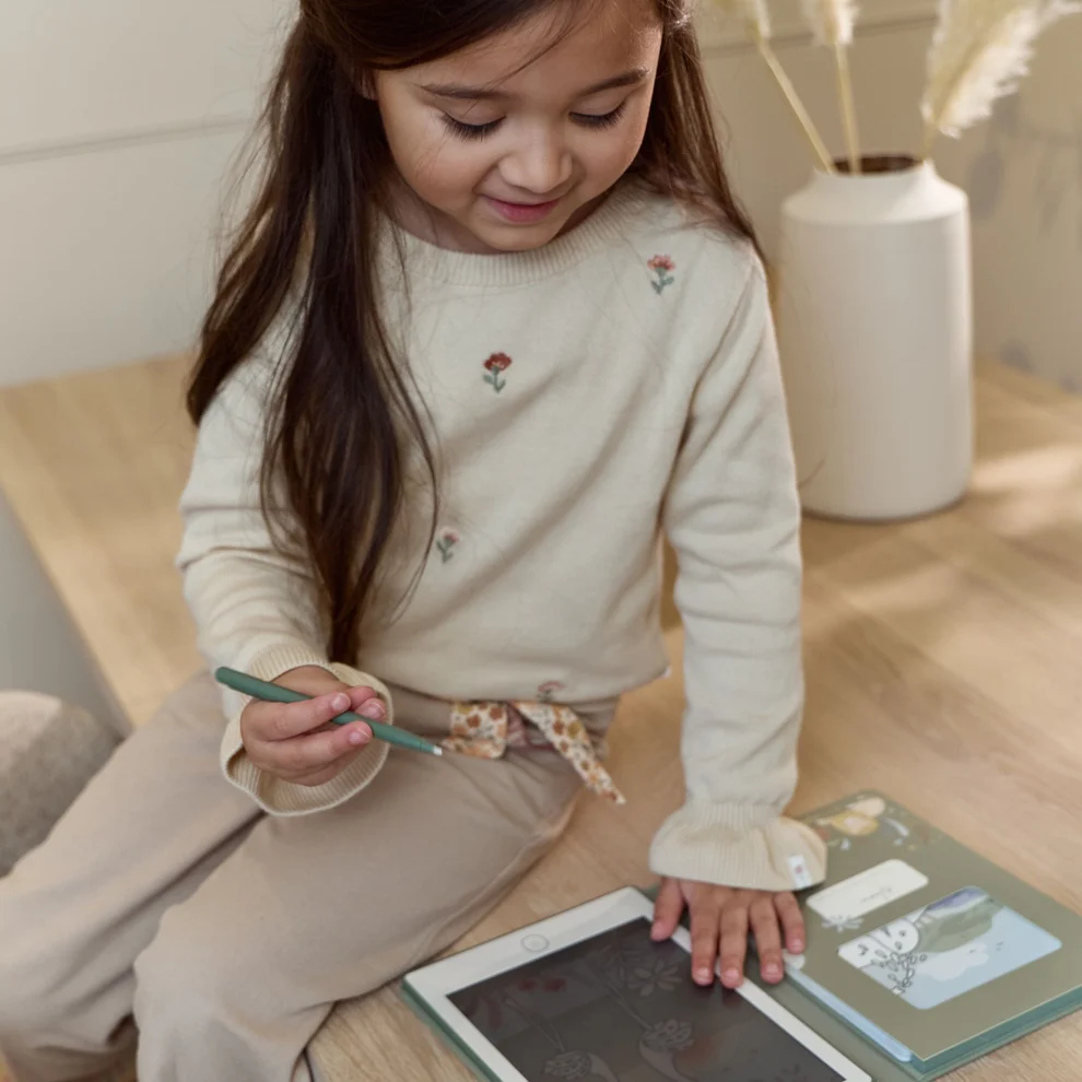 Menina com camisola creme bordada e calças castanhas claras a usar tablet em superfície de madeira.