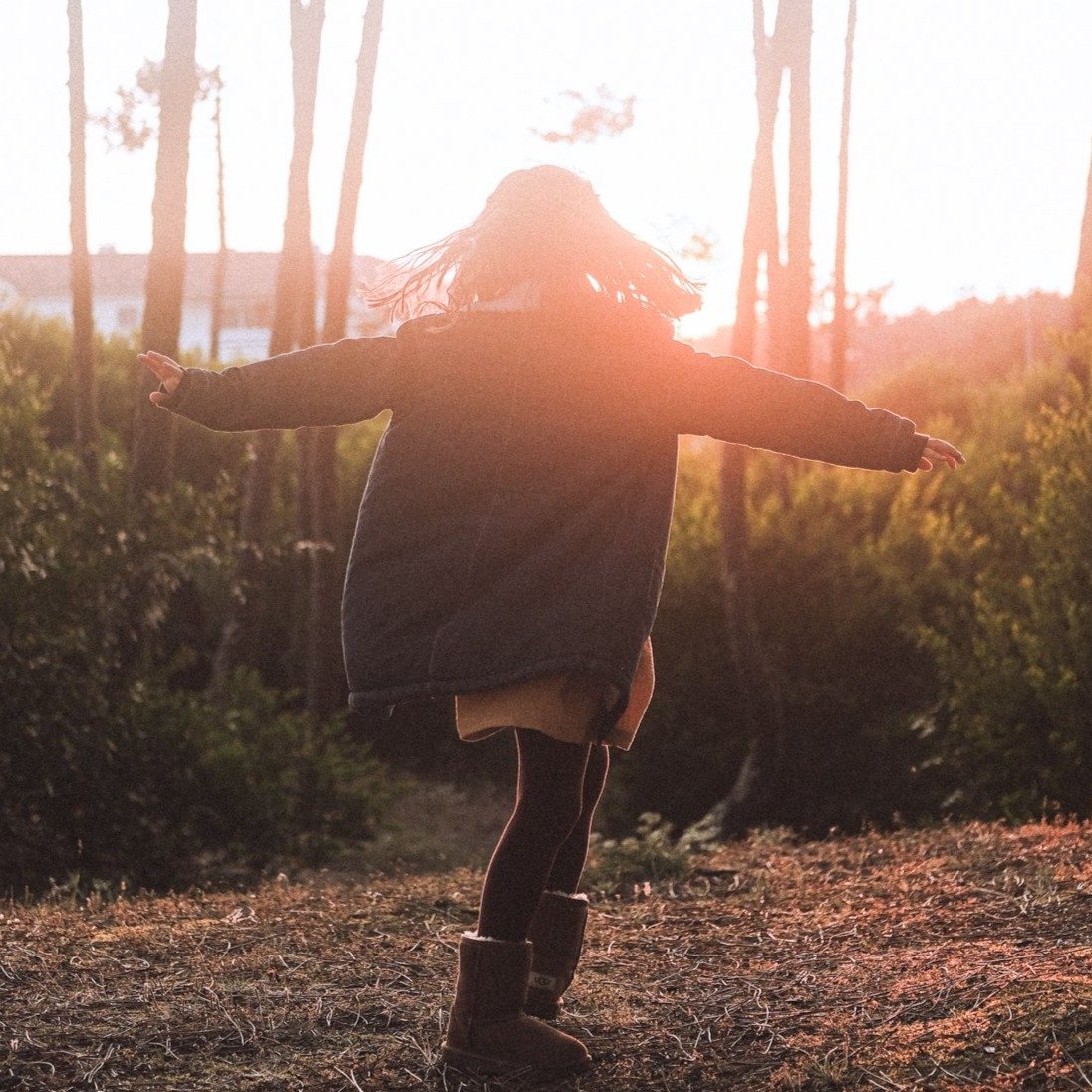 Menina de costas a girar num campo com botas, leggings e camisola escura.