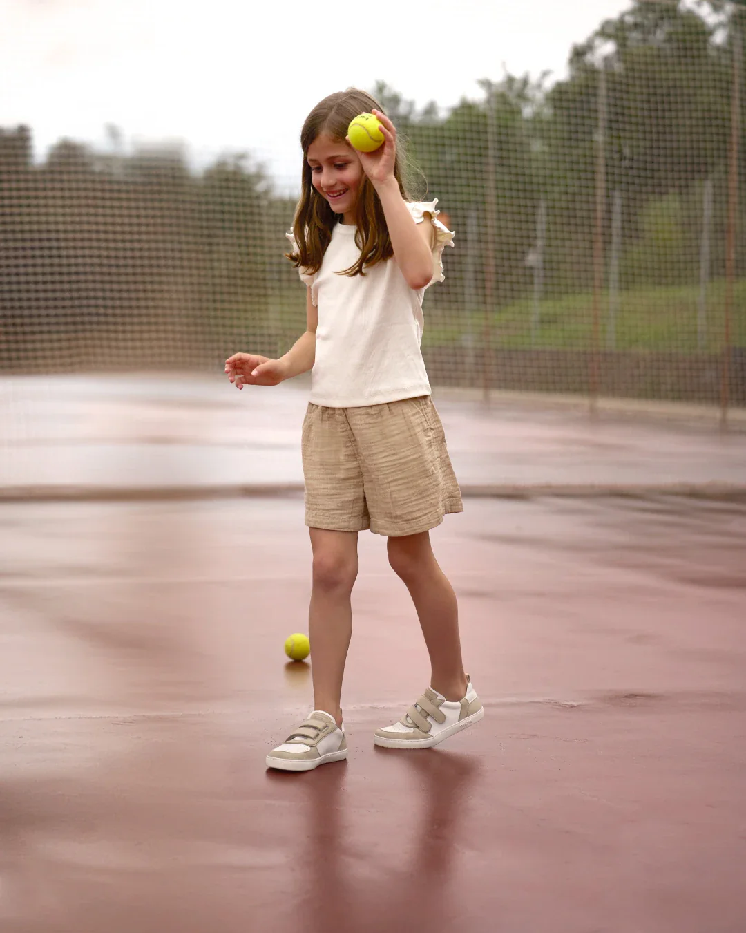 Criança a jogar ténis com roupa casual clara e ténis brancos numa quadra exterior molhada