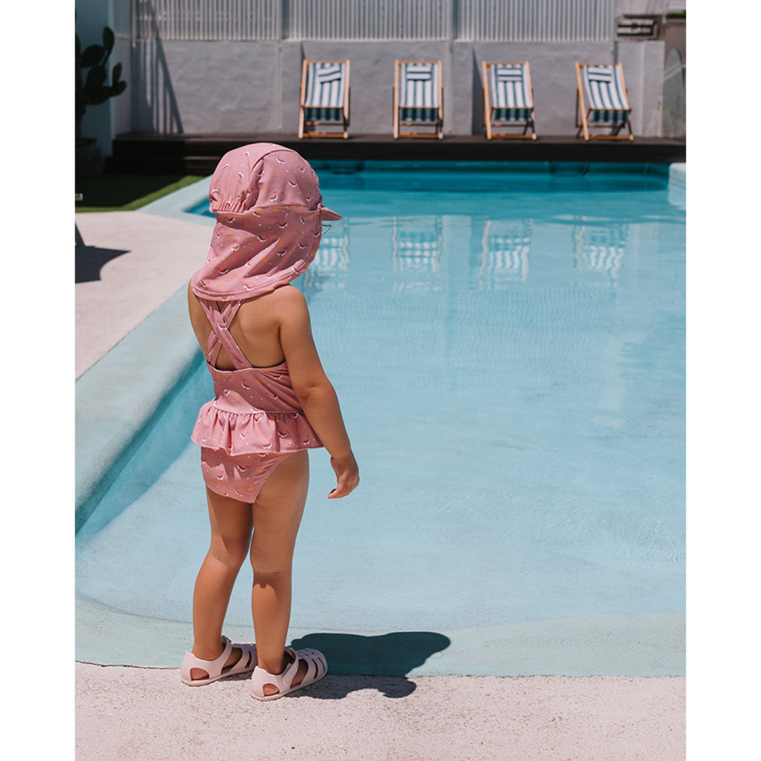 Criança de costas junto a uma piscina vestindo fato de banho cor de rosa com padrão e chapéu combinando.