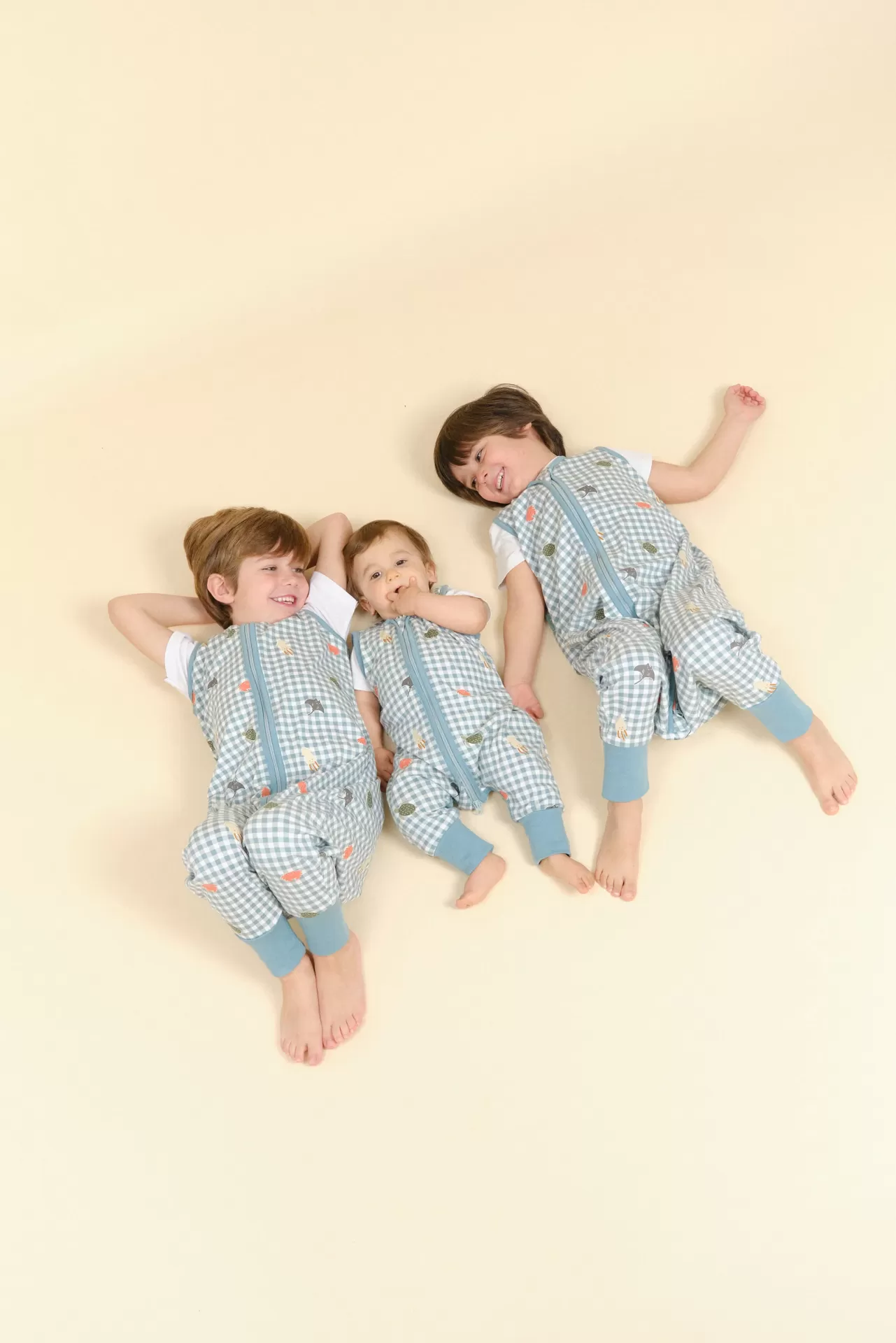 Três crianças deitadas usando pijamas xadrez azul e branco com estampas coloridas e punhos azul claro.