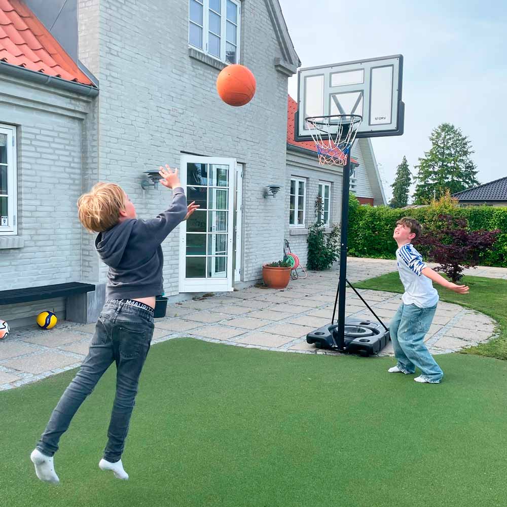 Mesa de basquetebol portátil com base preta num jardim exterior junto a uma casa cinza com telhado vermelho