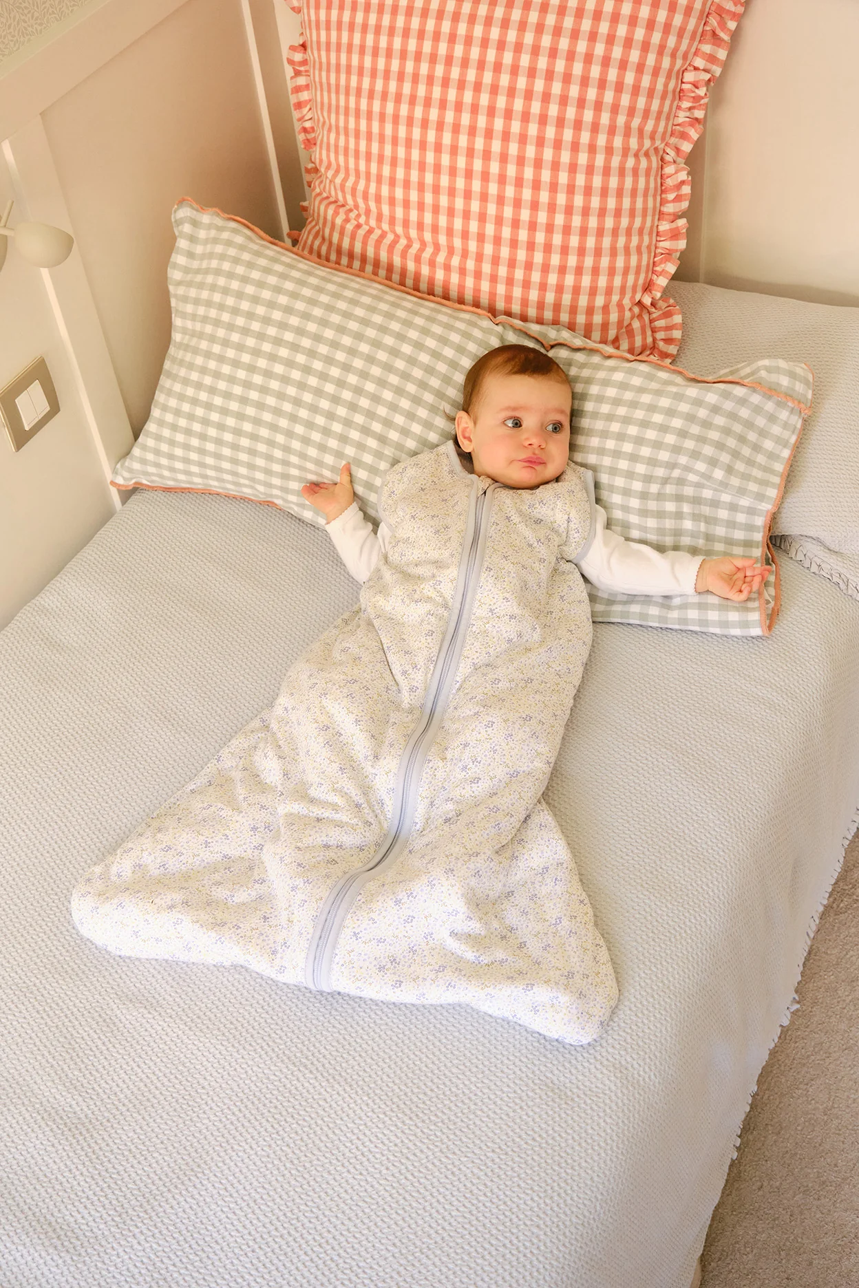 Bebé deitado numa cama com saco de dormir floral e almofadas vichy rosa e cinza.