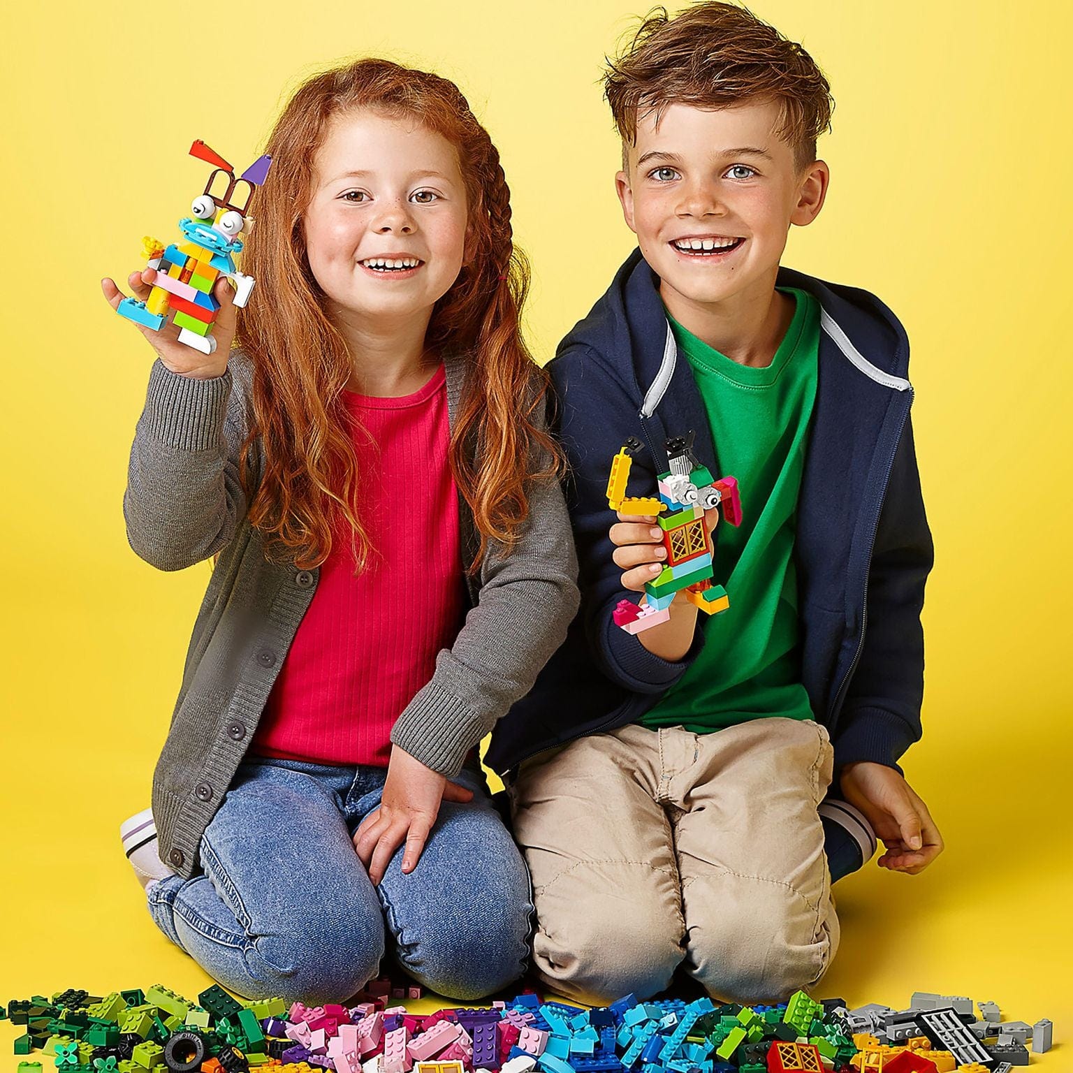 Duas crianças seguram figuras feitas com blocos de construção coloridos em fundo amarelo