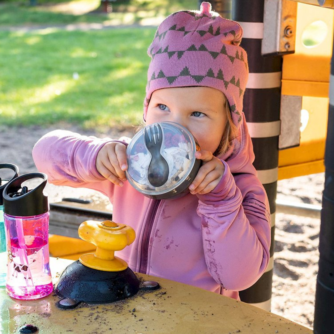 Menina com gorro cor de rosa a beber de recipiente plástico com talheres integrados, sentada numa mesa amarela de parque