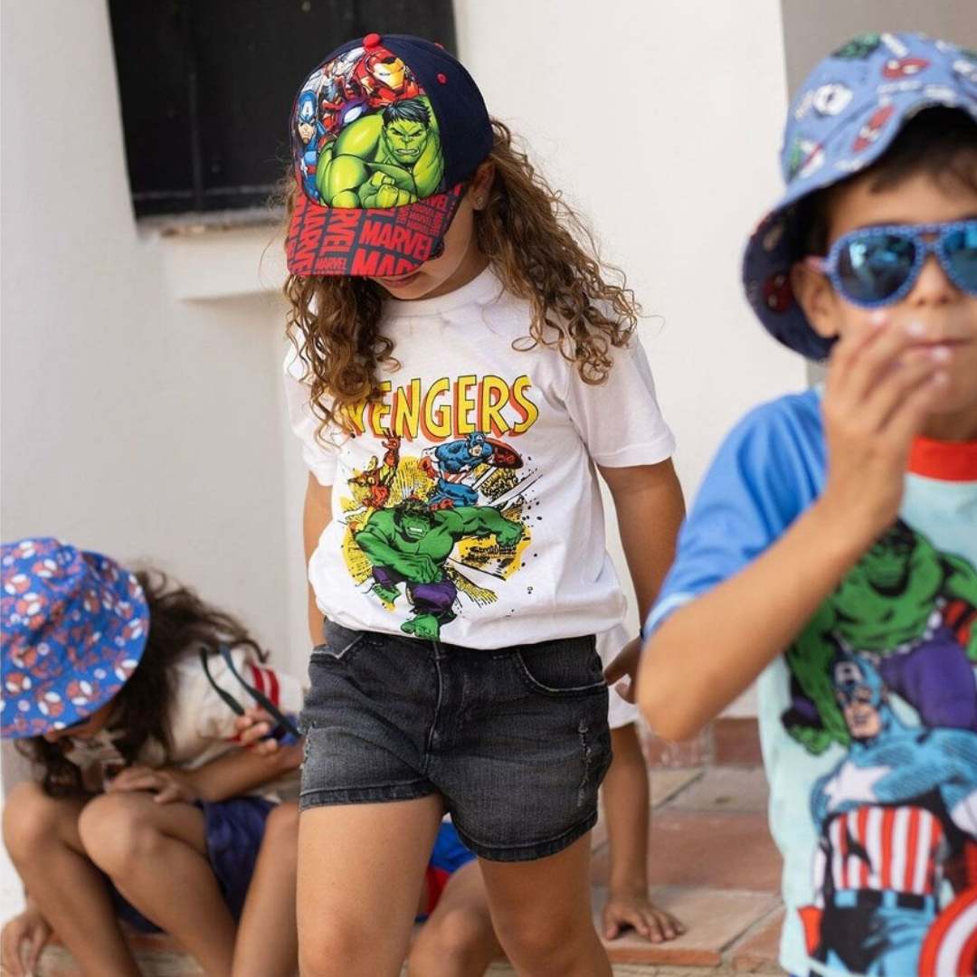 Criança com t-shirt dos Avengers e boné azul com Hulk, ao ar livre com outras crianças.