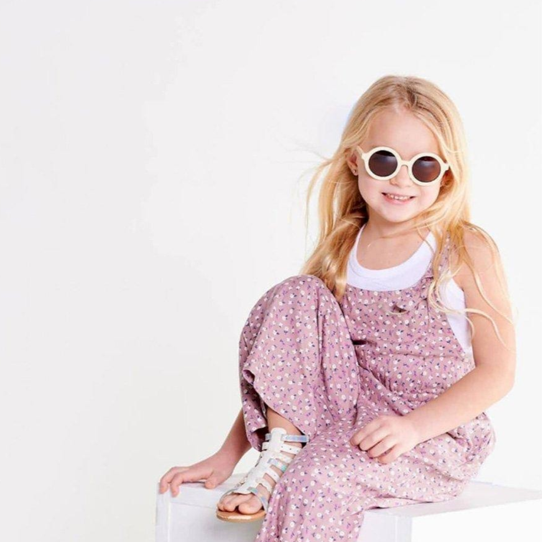 Criança loira sentada com macacão cor-de-rosa e óculos de sol brancos