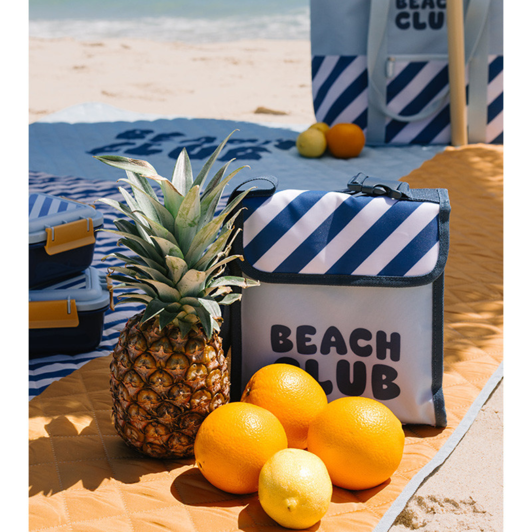 Saco térmico azul e branco com o texto BEACH CLUB numa praia com frutas à frente