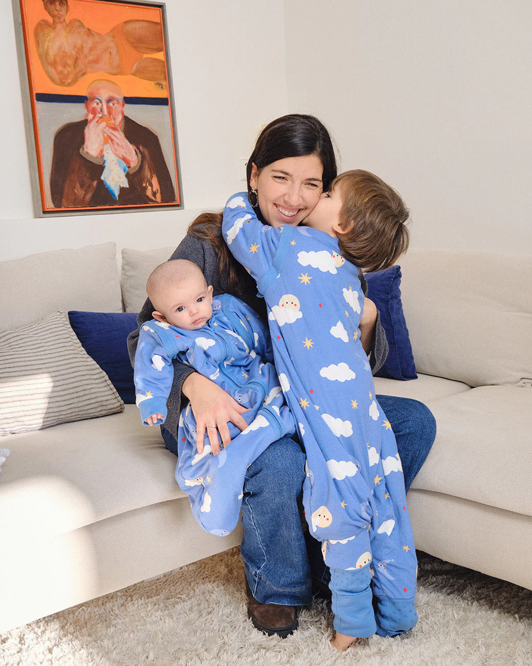 Pijamas azuis infantis com desenhos de nuvens e estrelas, bebé no colo e criança a abraçar mulher sentada em sofá bege.