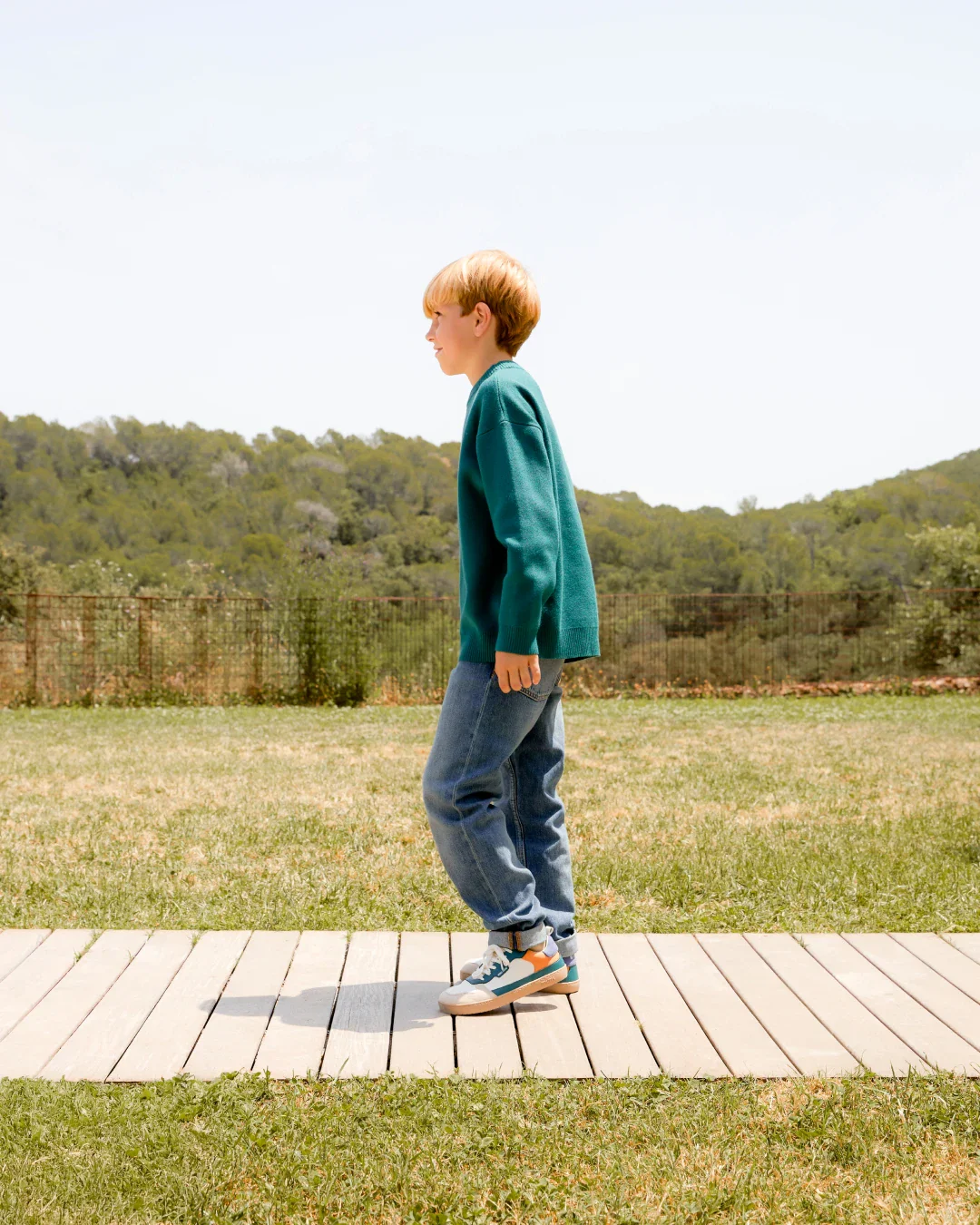 Menino com camisola verde-azulada, jeans e sapatilhas multicoloridas a caminhar numa passadeira de madeira ao ar livre.