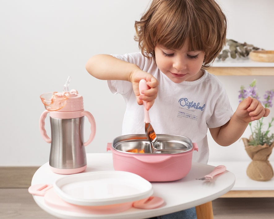 Conjunto de utensílios de alimentação infantil em rosa e branco numa mesa com uma criança a usar um garfo.