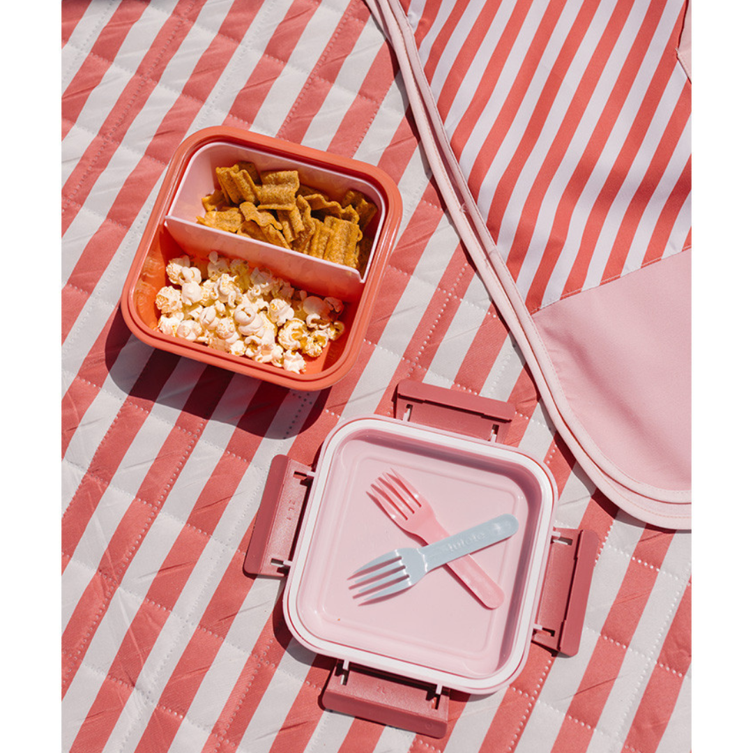 lancheira de plástico rosa e coral com comida e dois garfos sobre manta listrada cor de rosa e branca