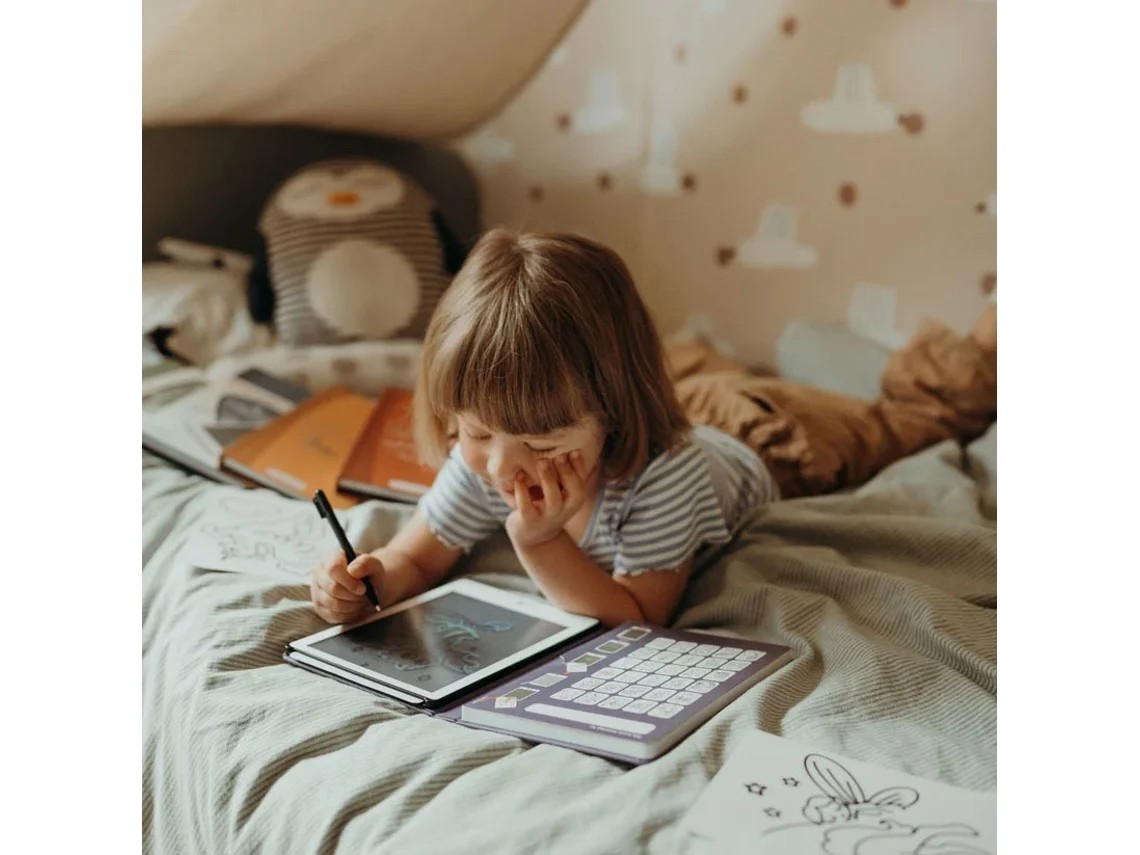 Menina a desenhar num tablet com teclado roxo, deitada numa cama com roupa cinzenta e almofadas castanhas num quarto infantil com papel de parede de nuvens.