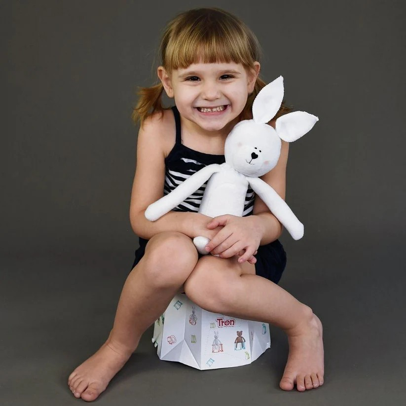 Menina sorridente segurando boneca de pano branca sentada em objeto hexagonal com texto 'Troh'