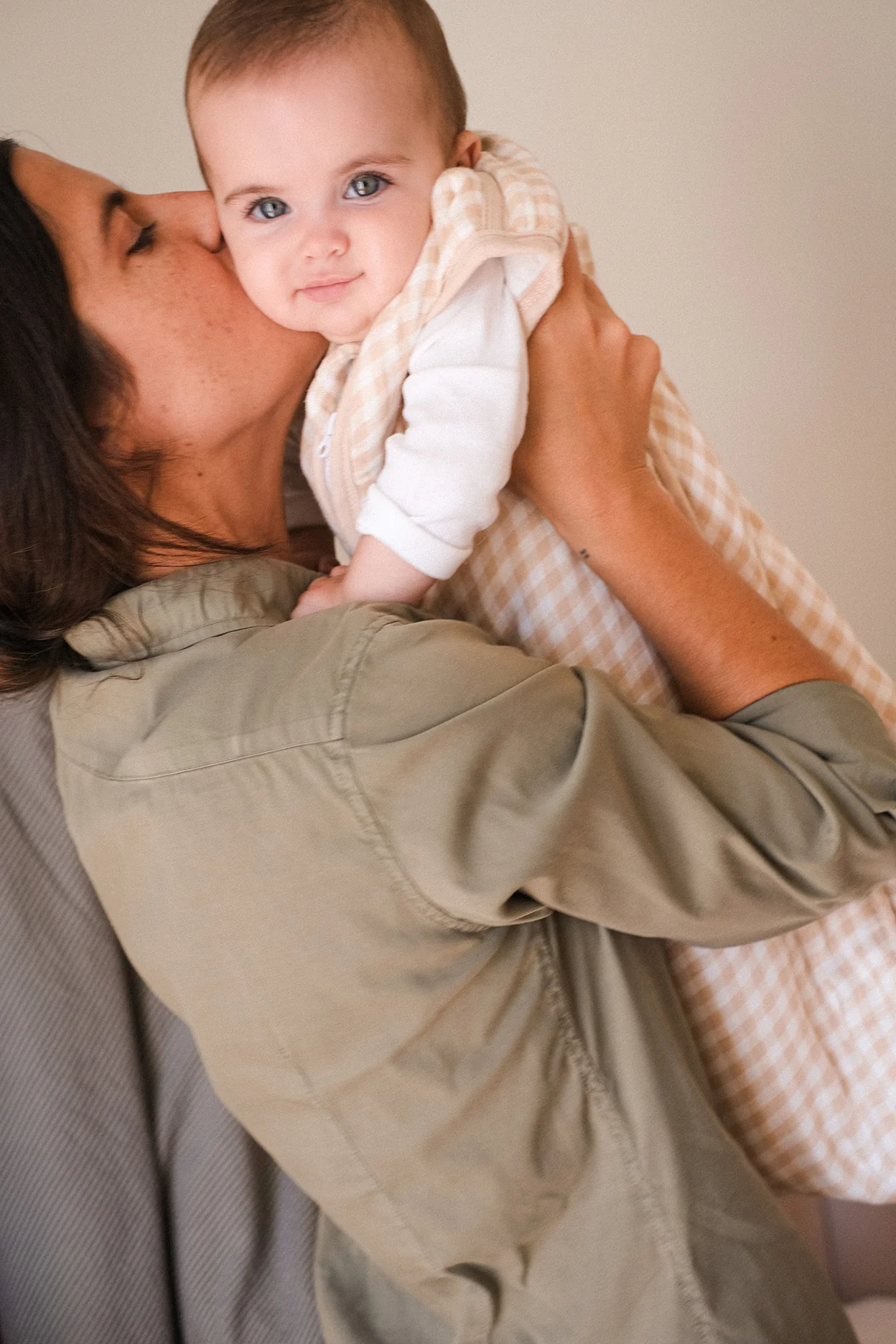 Bebé com colete xadrez bege e branco e body branco, abraçado por adulto com camisa verde-oliva