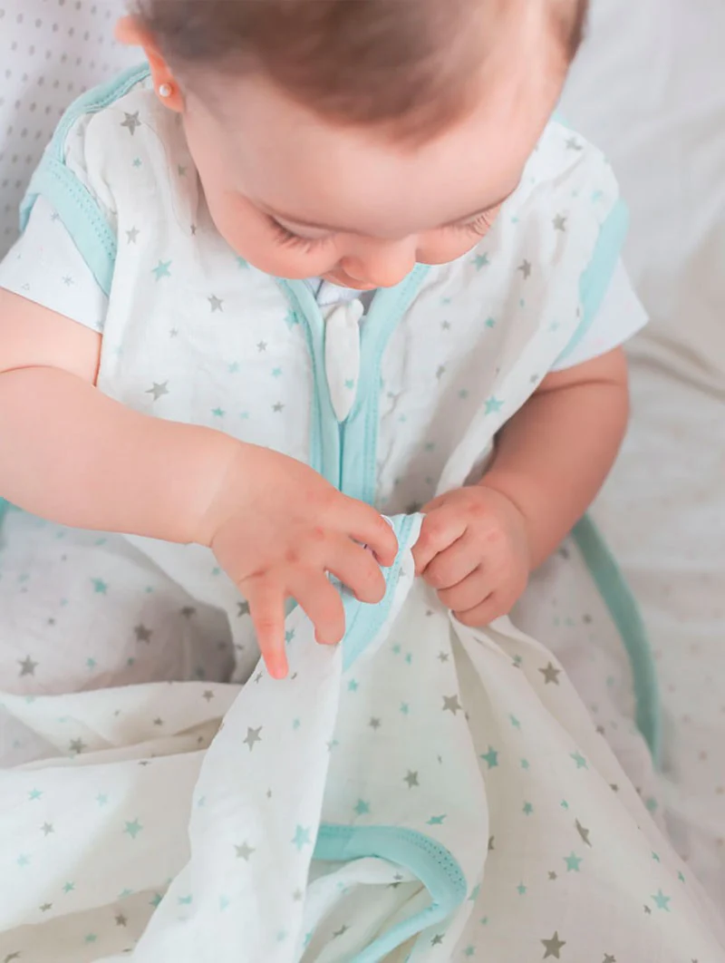 Bebé com pijama branco de estrelas cinzentas e azuis claras com bordas azuis claras sentado numa cama
