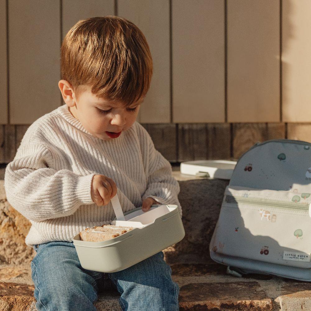 Criança com lancheira cinza e mochila cinza estampada sentada numa pedra