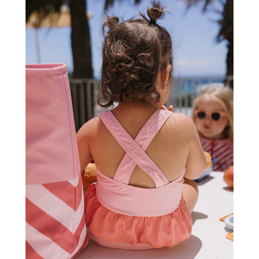 Menina de costas com fato de banho rosa claro e saia, sentada numa mesa branca ao ar livre