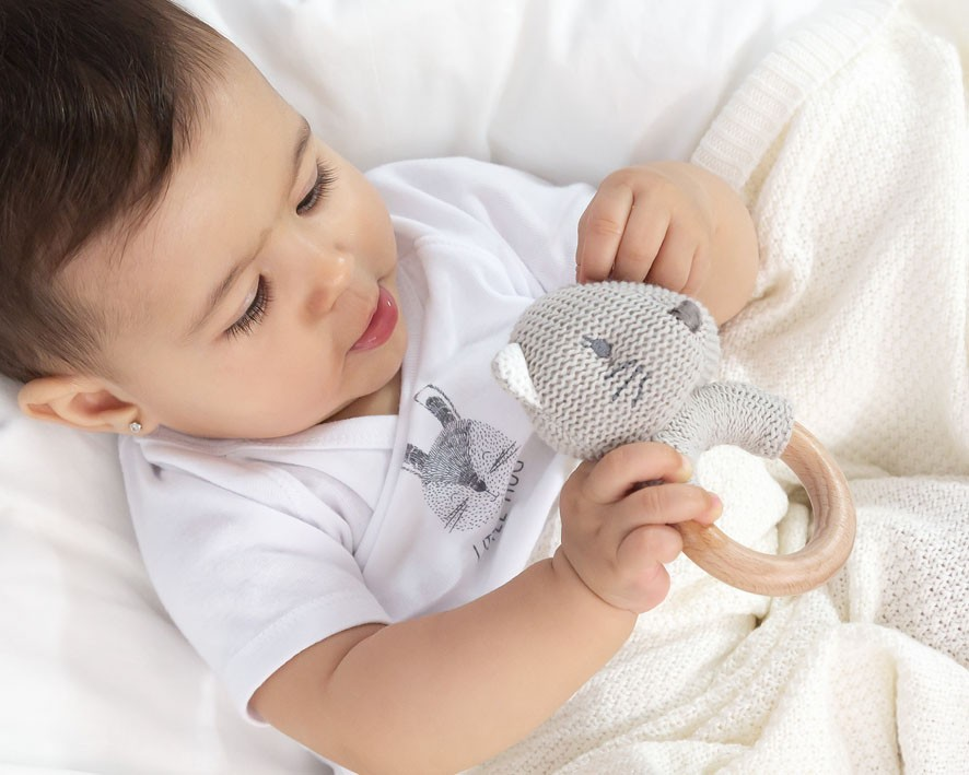 Bebé a segurar brinquedo de peluche cinzento com anel de madeira em cama branca