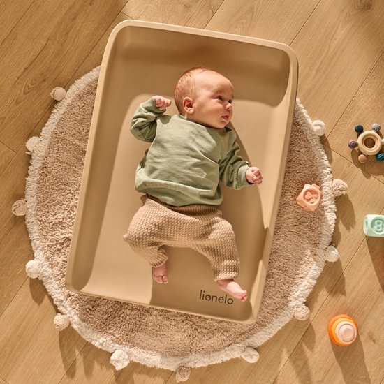 Bébé numa banheira bege sobre tapete creme com pompons em chão de madeira, brinquedos coloridos ao redor.