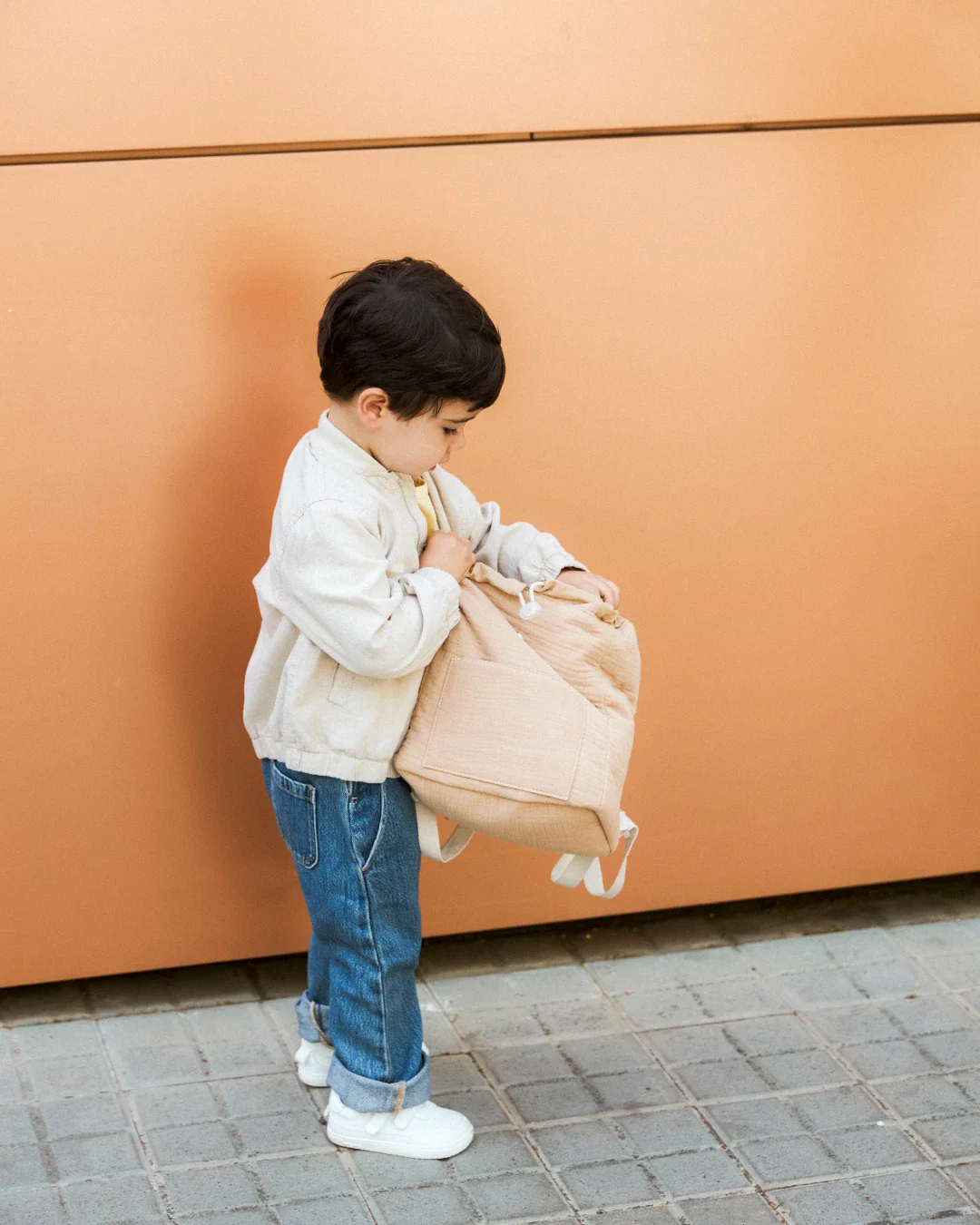 Criança com mochila bege de pé junto a parede laranja