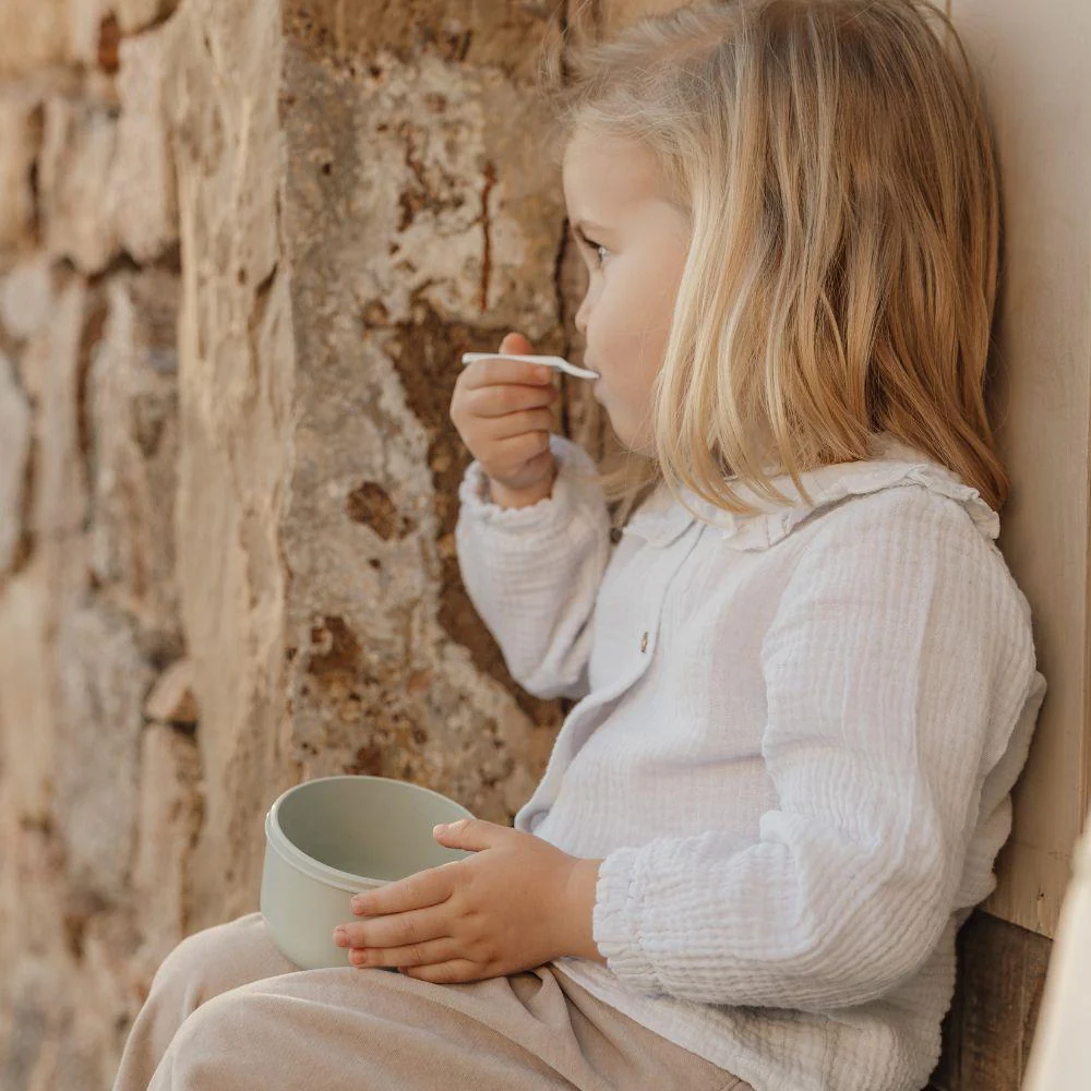 Menina loira com camisa branca segurando tigela cinza e colher branca junto a parede de pedra