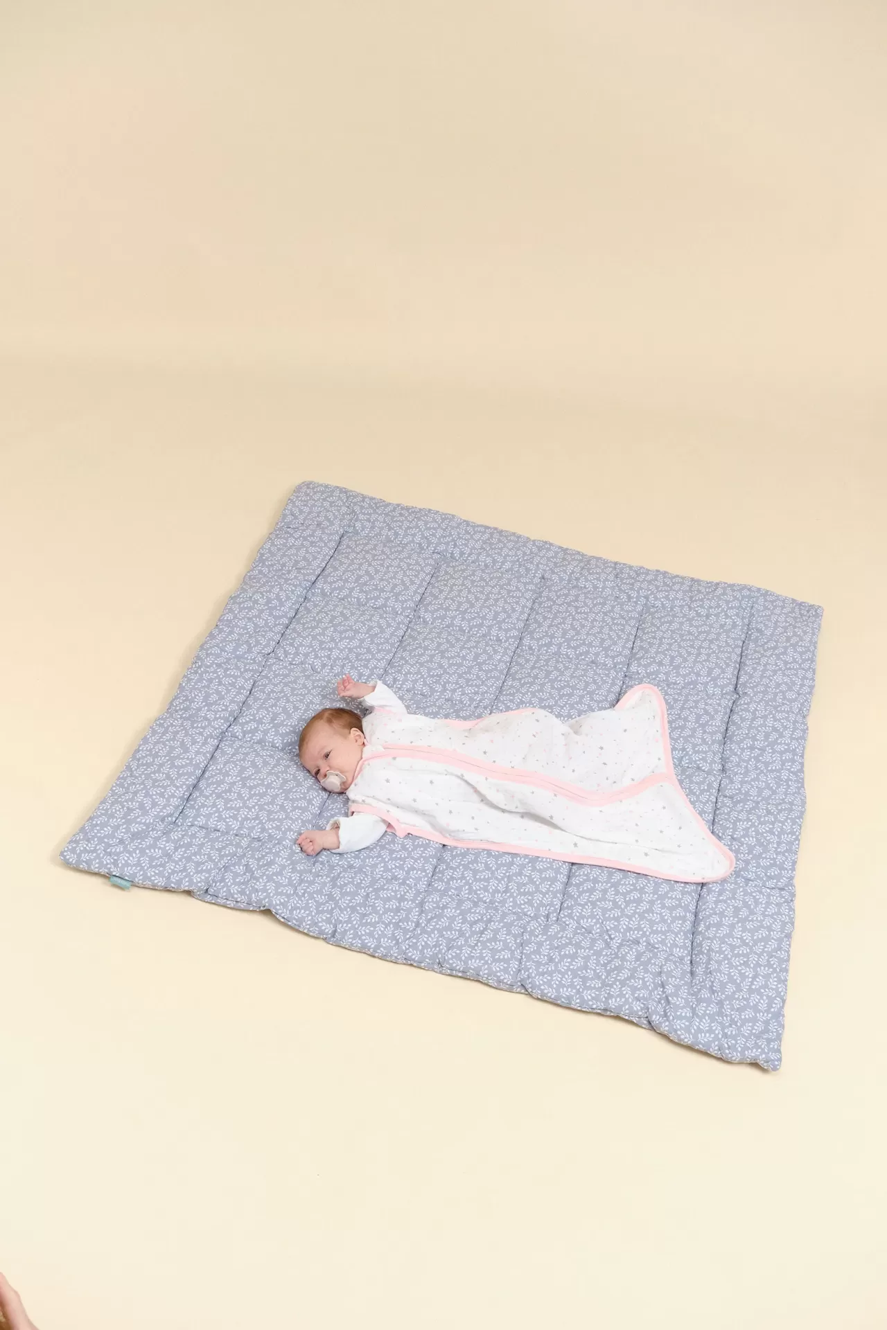 Bebé deitado sobre tapete almofadado cinza com padrão branco, saco de dormir branco com estrelas e bordas rosa.