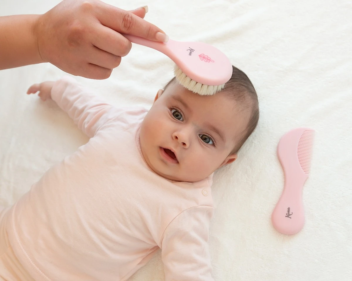 Bebê deitado sendo penteado com escova rosa sobre colchão branco macio