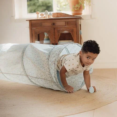 Criança a sair de túnel de brincar azul claro com padrão floral e segurando brinquedo azul