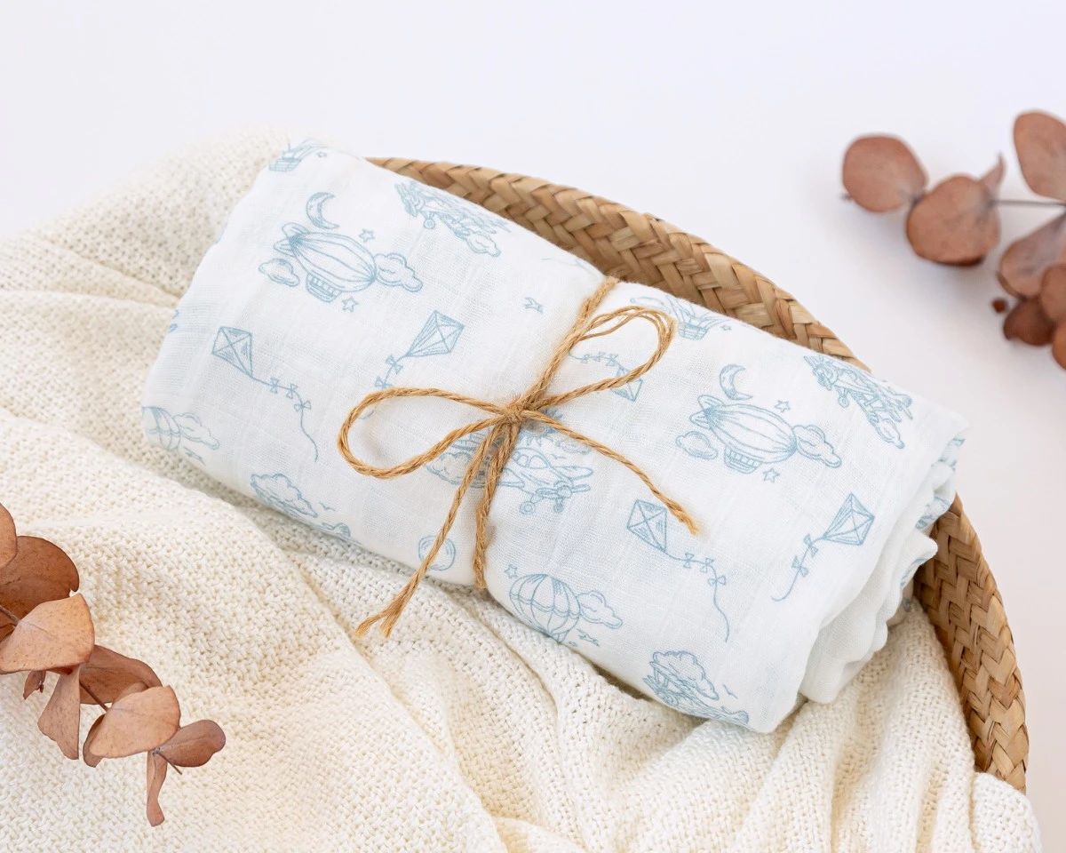 Fralda branca com padrão azul claro em cesto de palha com tecido branco e folhas secas ao lado